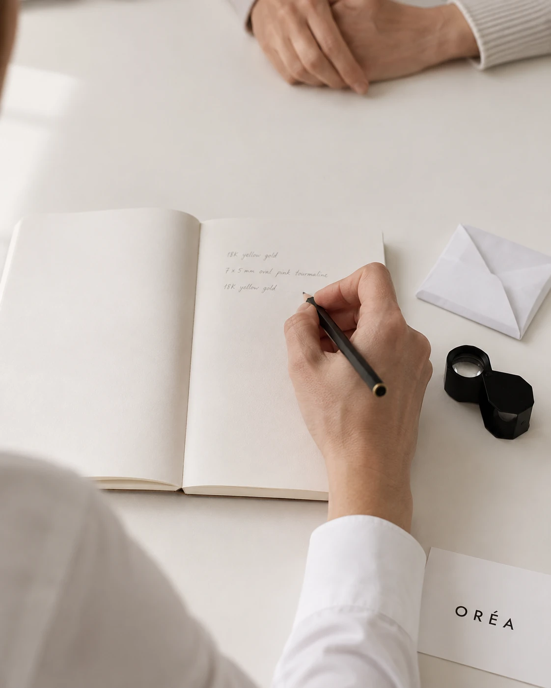 A hand writing notes in pencil in an open notebook across a pale table, a folded gem paper and a loupe set down beside it, the other person's hands resting still opposite