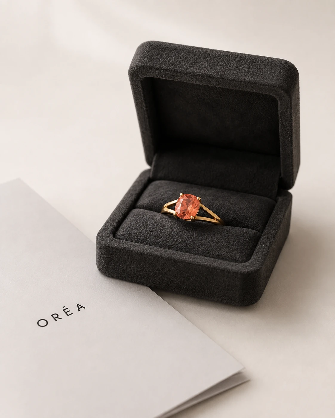 The finished gold ring, set with the orange pink cushion cut sapphire, in an open charcoal suede box with a folded sheet lying beside it