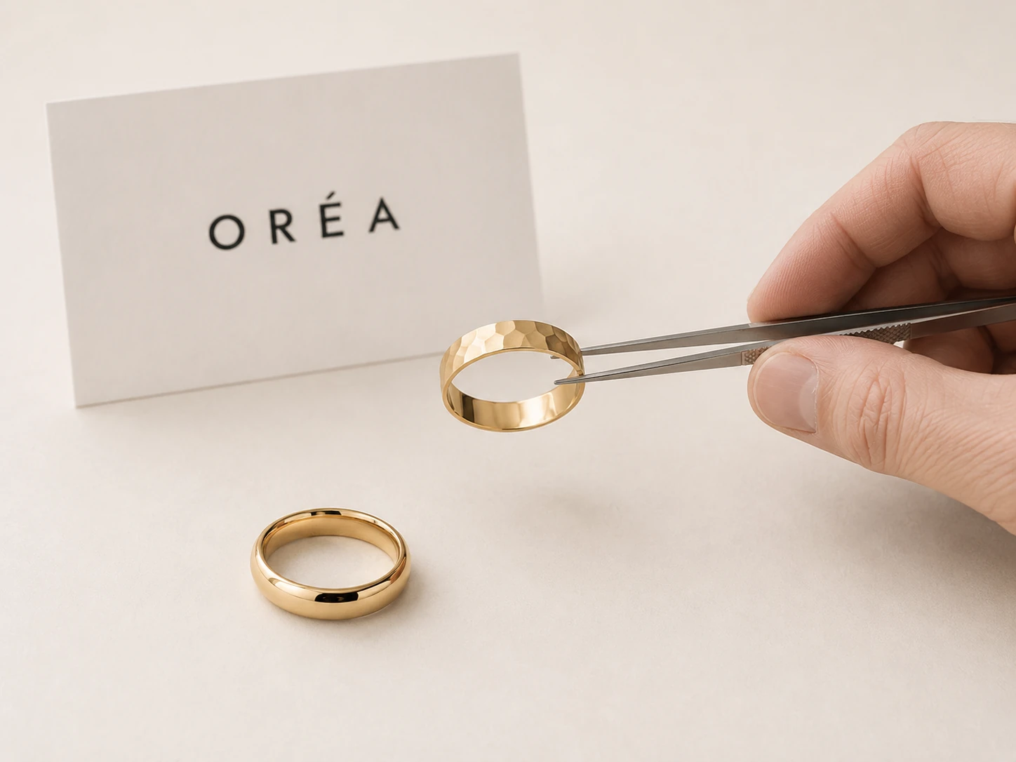 A hammer worked gold band held upright in steel tweezers, its facets catching the light, above the finished polished band of the same design lying on the sweep